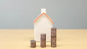 A small model house standing behind increasing stacks of British coins on a table.