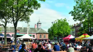 People sit at long wooden picnic tables in a sunny town square market.