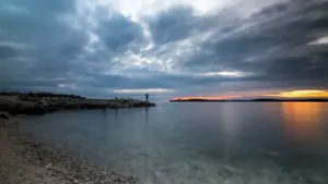 A solitary figure stands on a rocky shore overlooking a moody, expansive sea sunset.