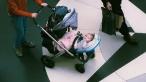 Parent pushing a baby in a stroller through a public indoor shopping area.