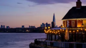 Illuminated riverside pub with the London city skyline at twilight.