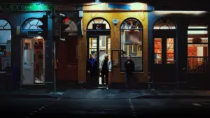 People standing outside illuminated evening storefronts on a city street at night.